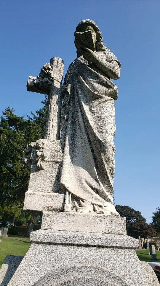 A carved granite statue of a woman holding a book and leaning on a cross, against a blue sky